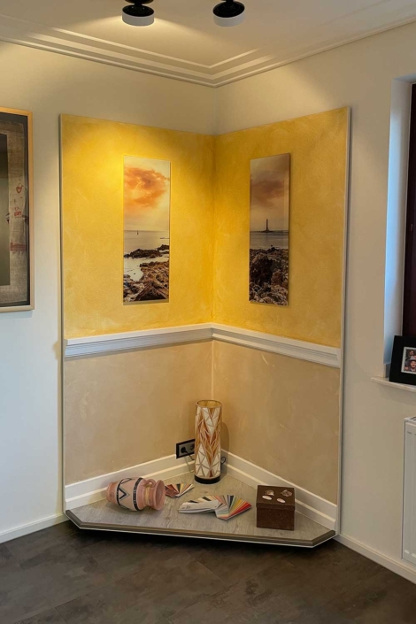 Gestaltete Showroom-Wandecke – Malerfachbetrieb Binder Dekorative Wandecke im Showroom des Malerfachbetriebs Binder mit gestalteten Wandflächen in Gelb-Beige, Bildern, Zierleiste und Podest mit Dekorationselementen