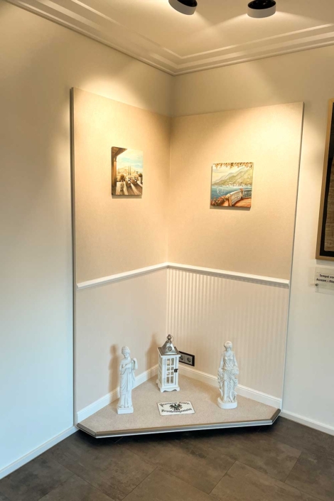 Showroom-Koje mit mediterraner Wandgestaltung – Malerfachbetrieb Binder Dekorativ gestaltete Showroom-Koje des Malerfachbetriebs Binder mit cremefarbener Wandstruktur, Zierleiste, mediterranen Bildern und klassischen Dekoobjekten